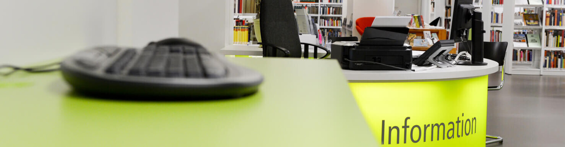 Linkerhand sieht man eine Tastatur im Vordergrund, rechterhand im Hintergrund sieht man die Infotheke der Bücherei Header_Infos_zur_Benutzung.jpg
