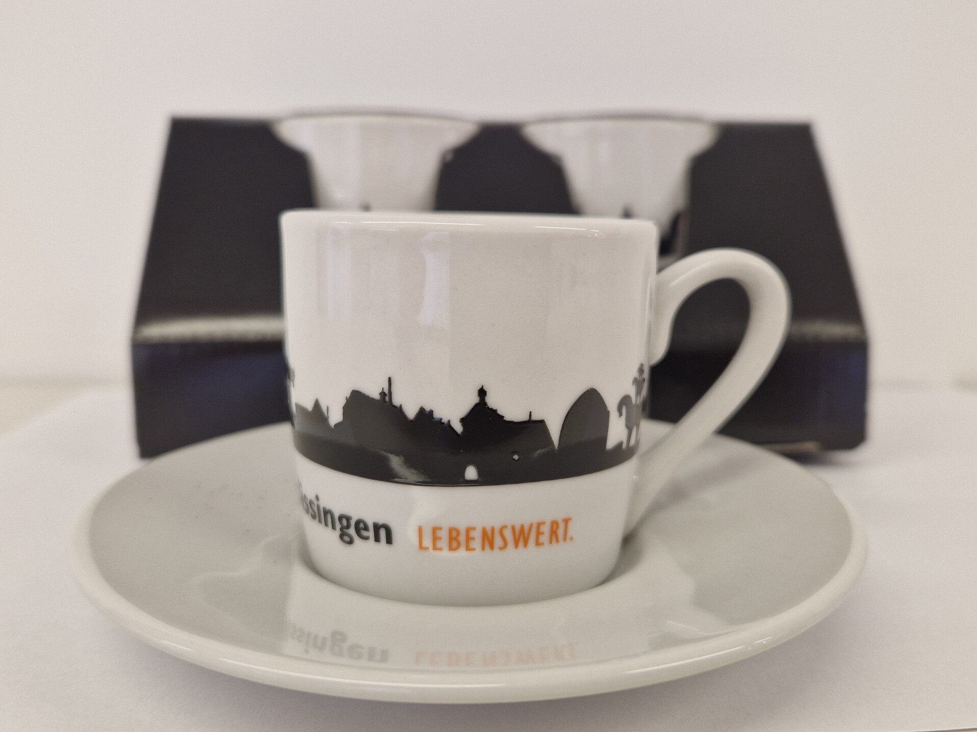 Eine weiße Espressotasse steht auf dem Unterteller. Auf der Tasse ist eine schwarze Stadtsilhouette zu sehen. 