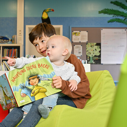 Ein Grundschulkind hat ein Kleinkind auf dem Schoß, zusammen schauen Sie sich ein Bilderbuch an Header_Kinder.jpg
