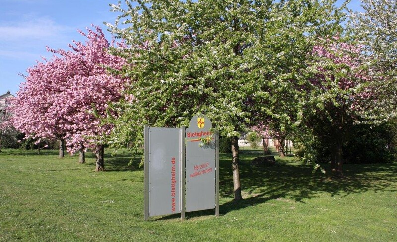 Willkommensschild auf einer Wiese vor blühenden Bäumen