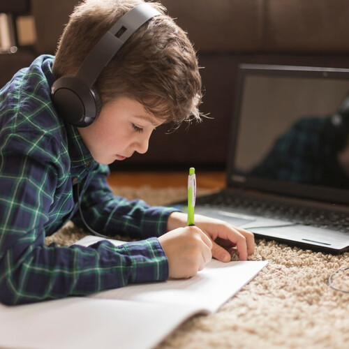 Lernender Junge auf dem Boden liegend, schreibt in ein Heft, während er Musik hört. Im Hintergrund sieht man einen geöffneten Laptop. Header_Grundschule.jpg