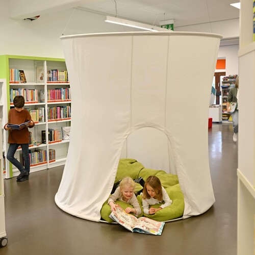 Einblick in die Kinderbücherei: Zu sehen sind Kinder die im Lesezelt auf Kissen liegen und lesen. Außerdem sieht man noch Kinder wie Sie Bücher in den Regal suchen. Header_Kita.jpg