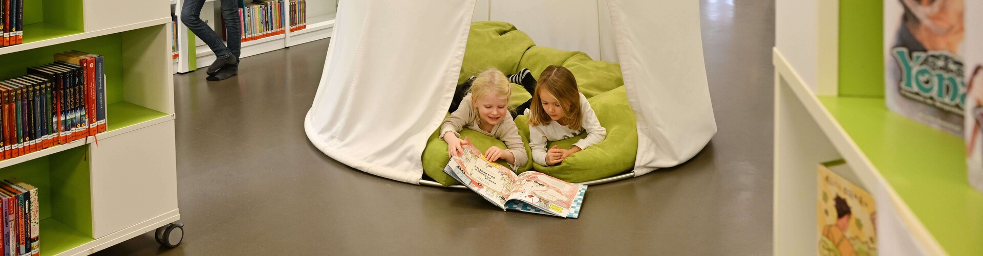 Einblick in die Kinderbücherei: Zu sehen sind Kinder die im Lesezelt auf Kissen liegen und lesen. Außerdem sieht man noch Kinder wie Sie Bücher in den Regal suchen. Header_Kita.jpg