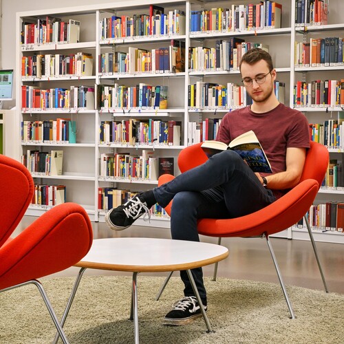 Im Vordergrund stehen zwei Sessel, auf einem davon sitzt ein junger Mann und liest ein Buch, im Hintergrund sind Bücherregale und eine junge Frau an einem Computer Header_Erwachsene.jpg