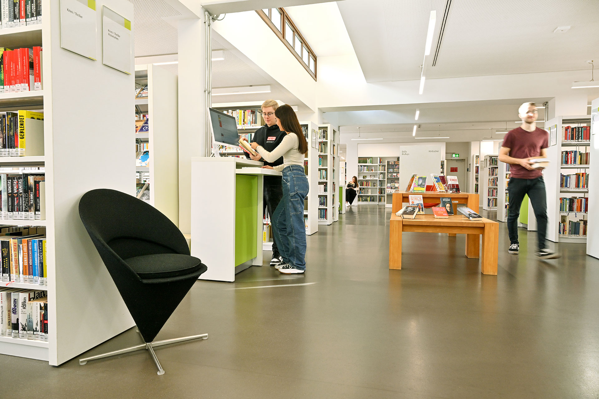 Bild der Büchereiräumlichkeiten, linkerhand sieht man Regale und einen Sessel und 2 Personen an einem Computer stehend, in der Mitte sieht man Ausstellungstische, eine weitere Person und Bücherregale Header_Startseite.jpg